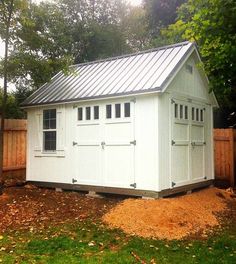 a white shed with a metal roof in the yard next to a fence and trees