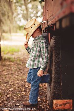 a young boy wearing a cowboy hat leaning on the side of a truck in the woods