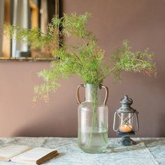 a vase filled with green plants sitting on top of a table next to an open book