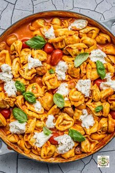 a skillet filled with pasta and vegetables on top of a stone floor next to a spoon