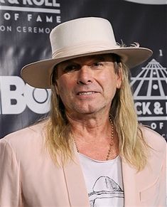 a man with long hair wearing a white hat and pink blazer standing in front of a sign