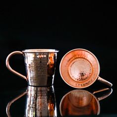 two copper colored cups sitting next to each other on a reflective counter top, with one cup in the foreground