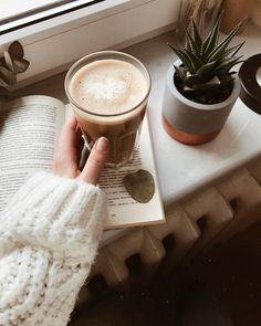 a person is holding a cup of coffee on top of an open book next to a potted plant