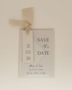 a white save the date card hanging on a wall with a ribbon attached to it