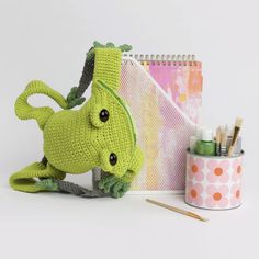 a crocheted green frog sitting next to a notebook and pencils