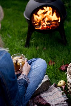 a woman sitting in front of an open fire pit holding a drink