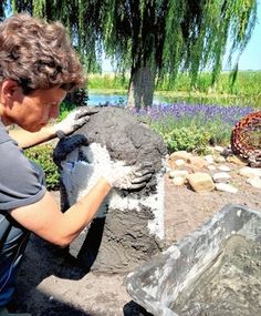 a woman is working on an object in the dirt near some water and trees with purple flowers behind her