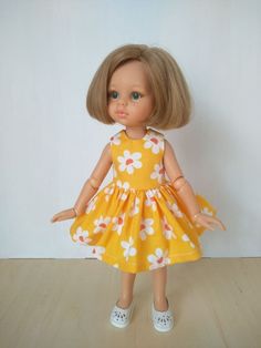 a doll with blonde hair wearing a yellow dress and white shoes, standing on a wooden floor