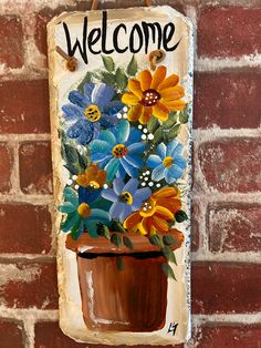 a welcome sign hanging on the side of a brick wall with flowers painted on it