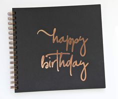 a spiral notebook with the words happy birthday written in copper foil on black paper next to a cup of coffee