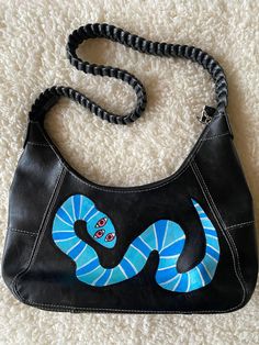 a black purse with a blue snake painted on the front and sides, sitting on a white carpet