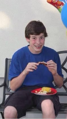 a young man sitting on a bench eating food with balloons in the air behind him