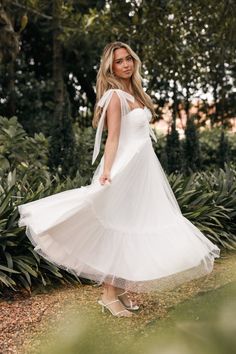 a woman in a white dress is posing for the camera with trees and bushes behind her