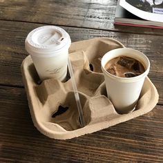 two cups of coffee sit in an egg carton on a table next to a book