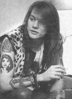 a man with long hair and tattoos on his arm sitting in front of a wall