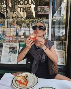 a woman sitting at a table eating food in front of a store window with glasses on her face