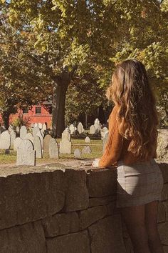 a woman standing next to a stone wall looking at the gravestones in front of her