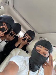 four people wearing black covering their faces in the back seat of a car with one person holding up his hand