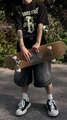 a young man holding a skateboard in his hands while standing on the street with trees behind him