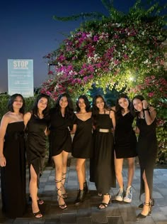 four women in black dresses posing for a photo at night with purple flowers behind them