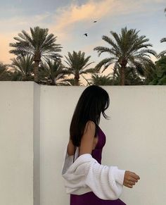 a woman in a purple dress leaning against a white wall with palm trees behind her