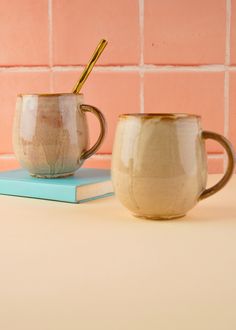 two coffee mugs sitting next to each other on top of a book near a pink brick wall