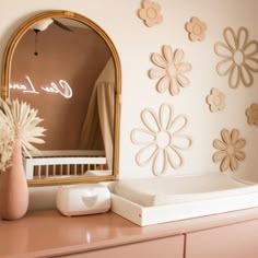 a dresser with a mirror, vase and flowers on it in front of a pink wall