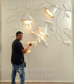 a man standing in front of a white wall with flowers on it and lights hanging from the ceiling