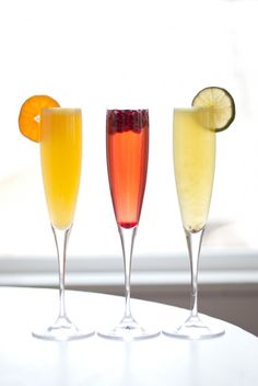 three glasses filled with different types of drinks on top of a white table next to a window