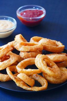 some onion rings are on a black plate with dipping sauces in bowls behind them