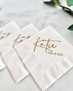 three white napkins with gold foil lettering on them and greenery in the background