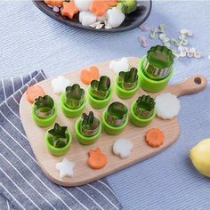 a wooden cutting board topped with green cups filled with carrots and broccoli