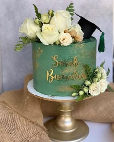 a green graduation cake with white flowers and greenery sits on top of a gold pedestal