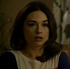 a woman with dark hair and blue eyes looks at the camera in a dimly lit room