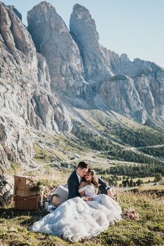 a bride and groom sitting on the grass in front of mountains