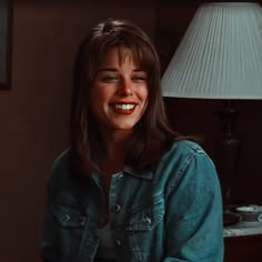 a woman sitting in front of a lamp smiling at the camera while wearing a jean jacket