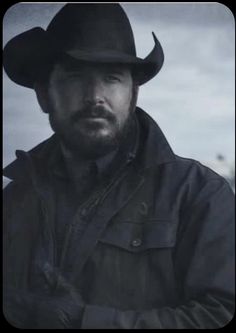 a man with a beard wearing a black cowboy hat and leather jacket standing in front of a cloudy sky