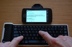 a person using a cell phone next to a keyboard on a wooden table with an electronic device