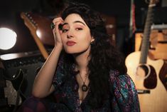 a woman with long hair sitting in front of guitars and holding her hand up to her face