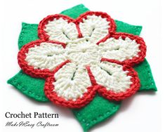 a crocheted flower with red and white petals on green leaves, sitting on a white surface