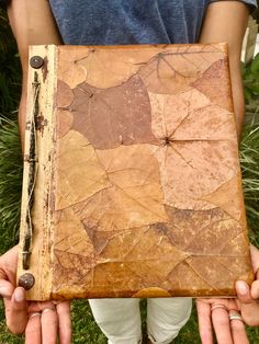 a person holding an old book with leaves on it in front of some grass and bushes