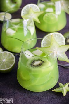 two glasses filled with green liquid and sliced kiwis on the rim, surrounded by lime slices