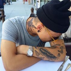 a man sitting at a table with tattoos on his face and arm, looking down