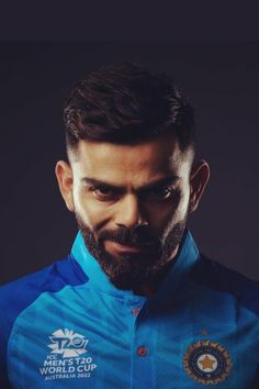 a man with a beard wearing a blue shirt and looking at the camera while standing in front of a dark background