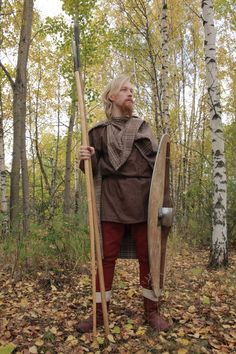 a woman dressed in medieval clothing holding a shield and spear standing in the woods with leaves on the ground