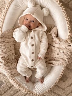 a baby in a white outfit laying on top of a blanket and wearing a hat