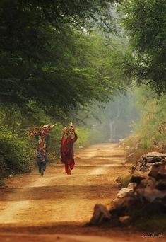 two people walking down a dirt road with baskets on their heads and trees in the background