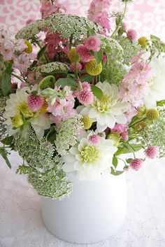 a white vase filled with lots of pink and white flowers on a tablecloth covered surface