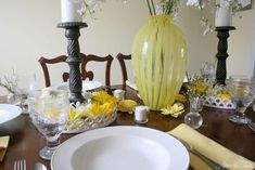 a dining room table is set with yellow flowers and white plates, silverware, and candlesticks