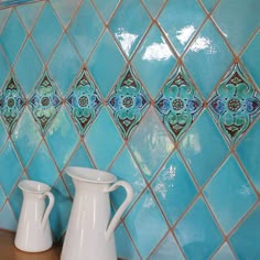 two white vases sitting next to each other on a wooden table in front of a blue tiled wall